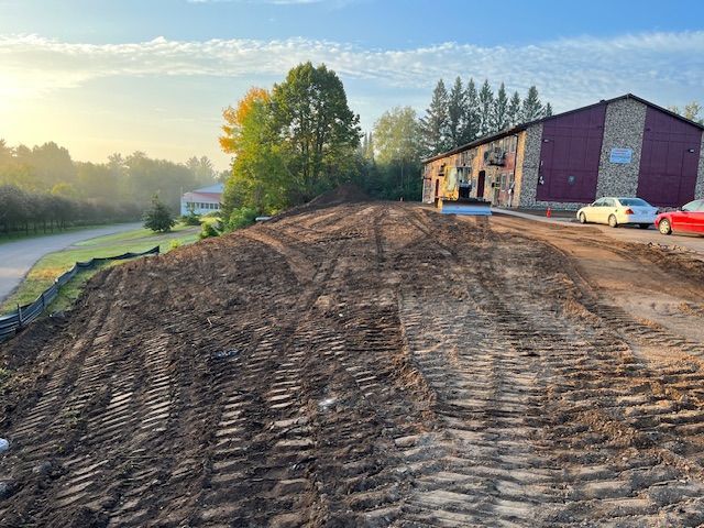 Dirt pile in front of a stone building with two parked cars. Trees and a residential building in the background.