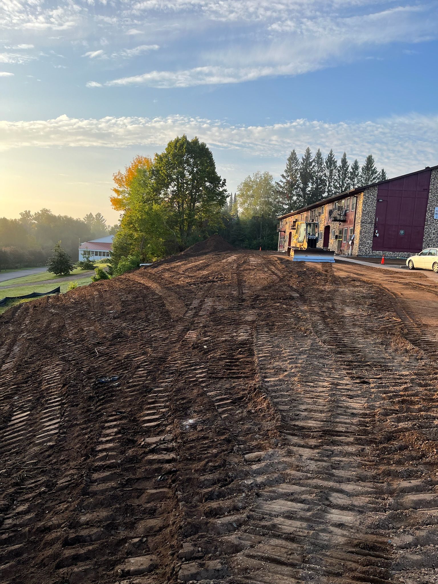 Dirt mound in foreground; building with red wall and trees in background; sky with sunlight.