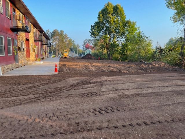Dirt ground with tire tracks in front of a red building with a new concrete sidewalk, trees in background.