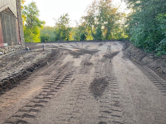 Dirt driveway with heavy tire tracks, possibly graded. Trees and building in the background.