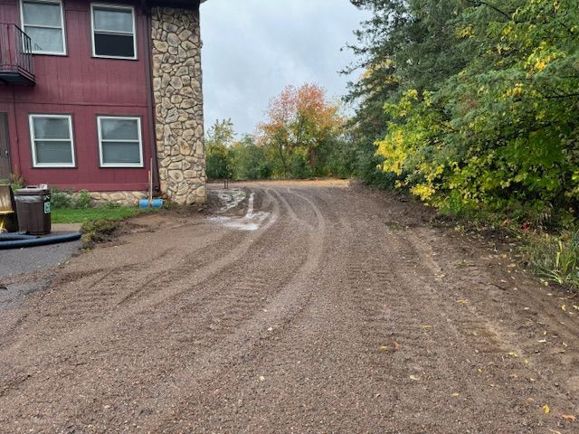 Dirt driveway with tire tracks leading to a house, surrounded by trees and a brownstone column.