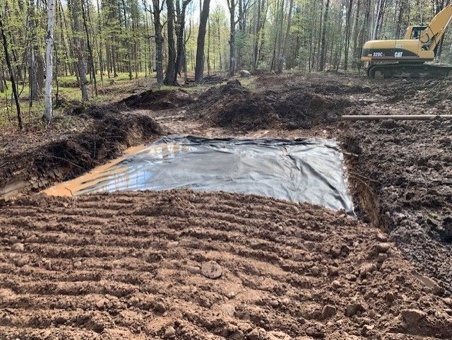 Excavation site with tarp-lined pit; backhoe in background, muddy terrain, and surrounding forest.