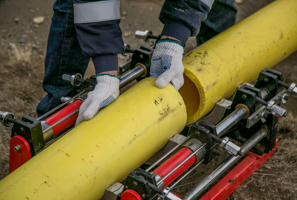 A worker wearing gloves uses a specialized alignment machine to join two sections of yellow polyethylene pipe.