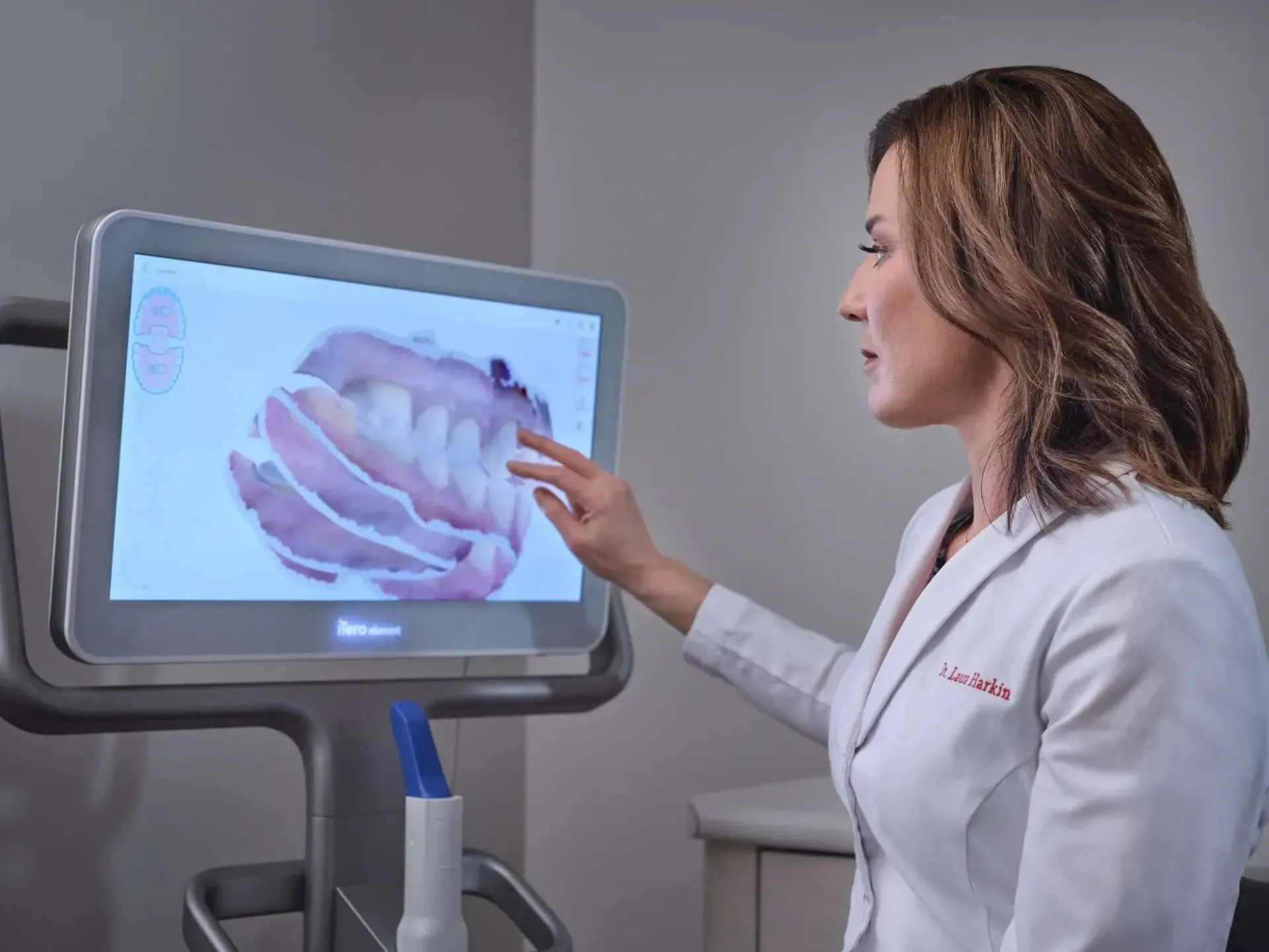Clinician in white coat pointing at a touchscreen display showing a dental image in a medical office