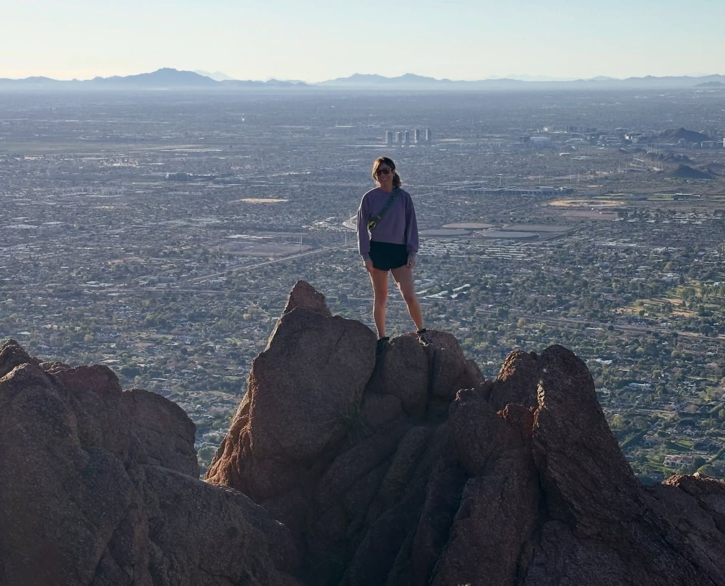 Hiker standing on rocky peak overlooking a vast cityscape at sunrise.