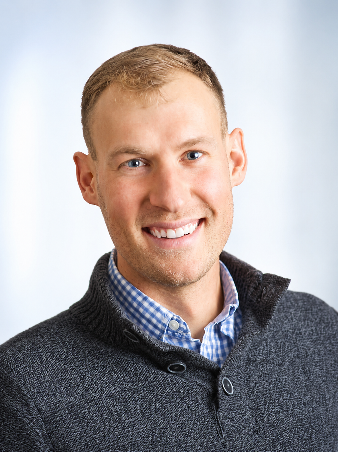 Smiling man in a blue checkered shirt and dark sweater against a light background