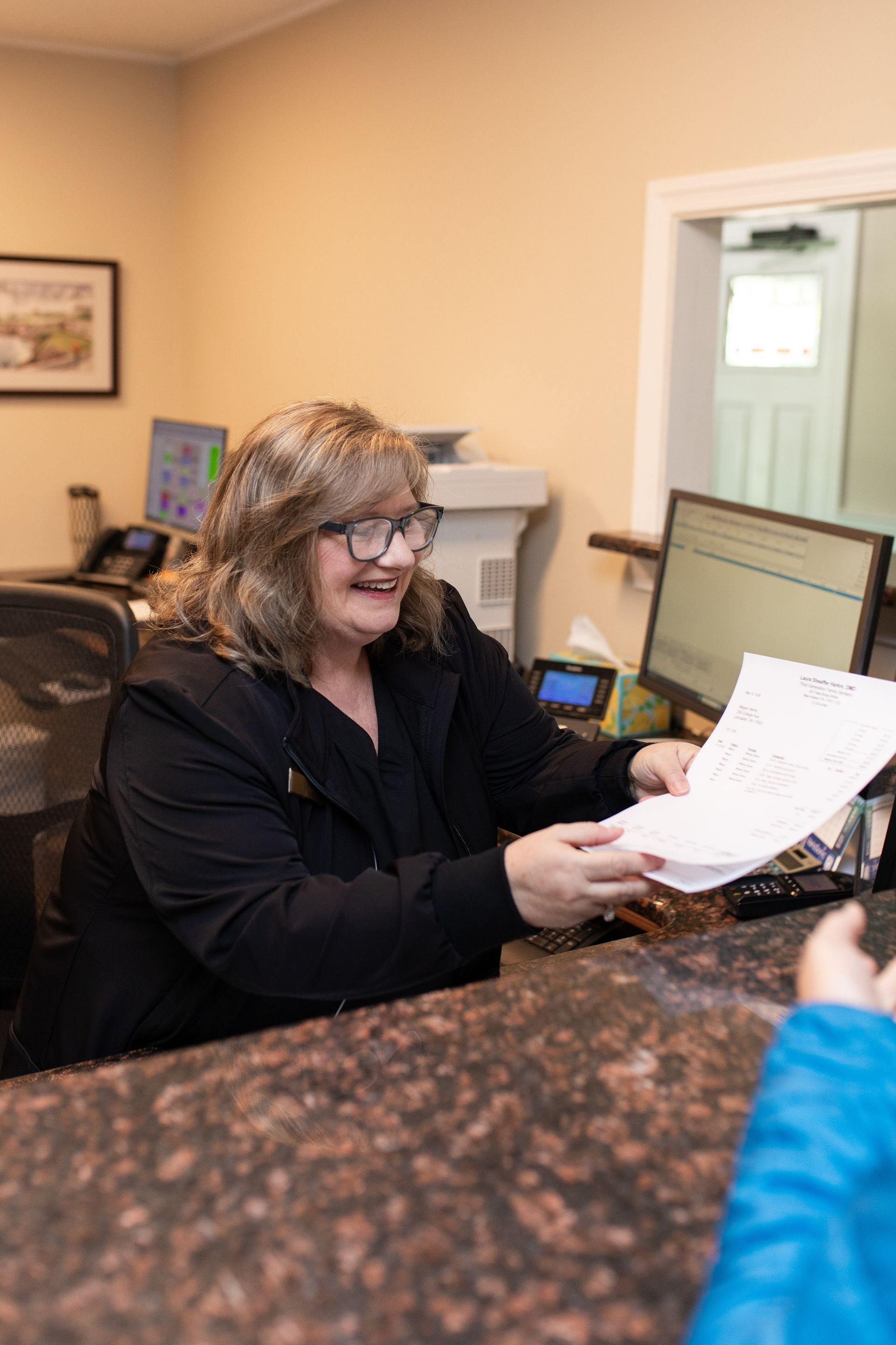 Woman at a desk in an office, reviewing papers with a printer and computer nearby.