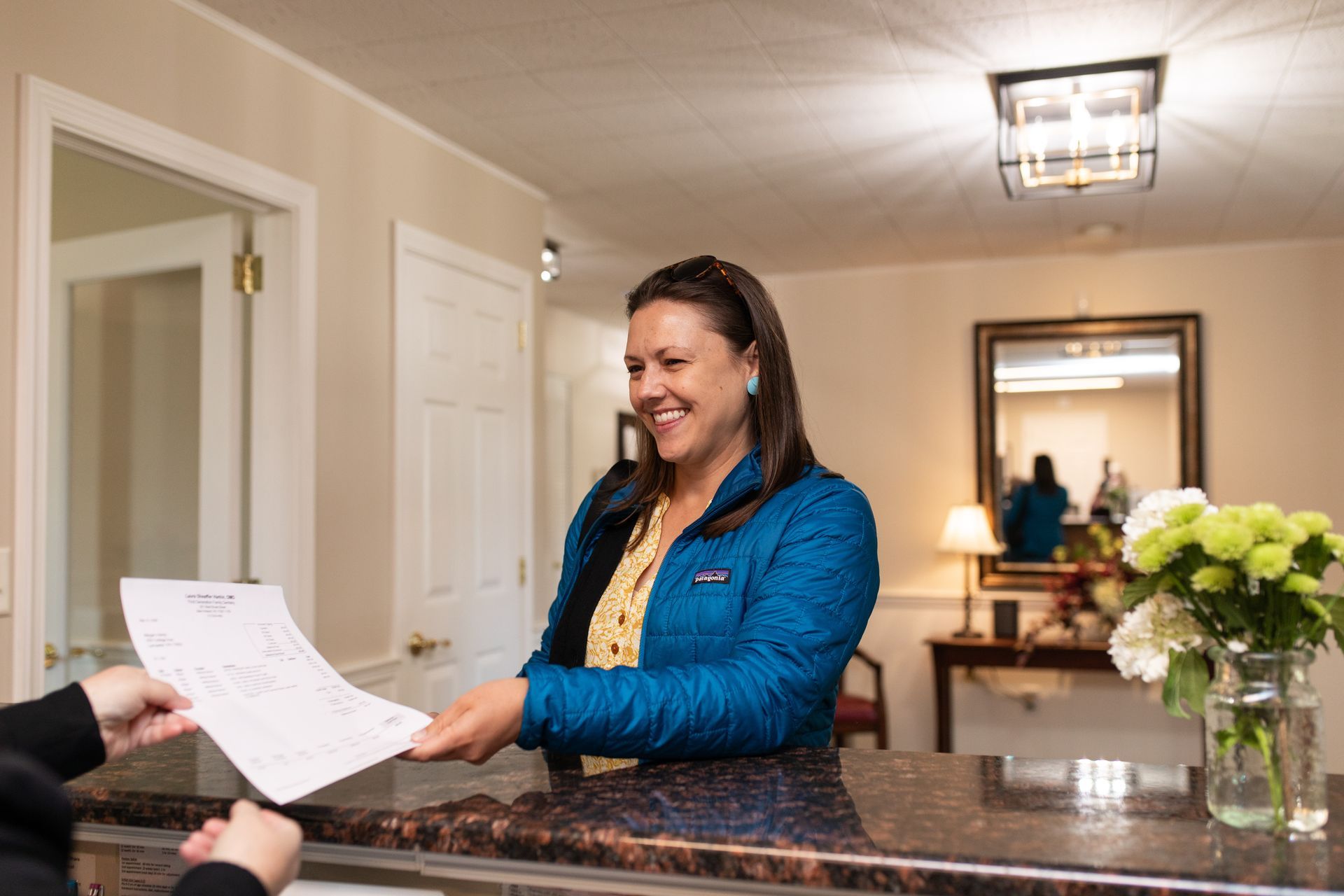 Woman at a hotel reception desk handing paperwork to a guest, smiling.