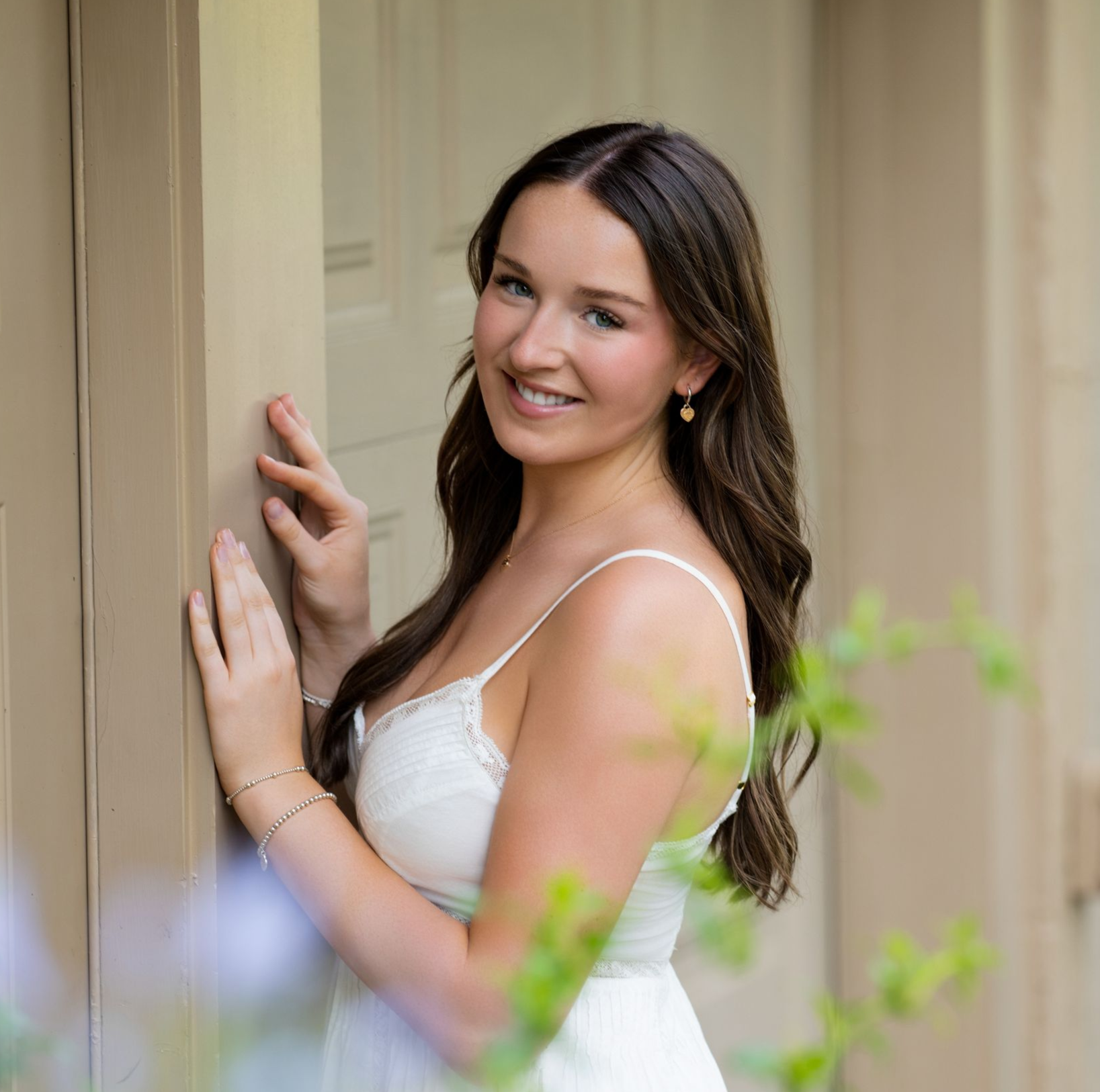 Woman in a white dress smiling by a doorway, with soft greenery in the foreground.