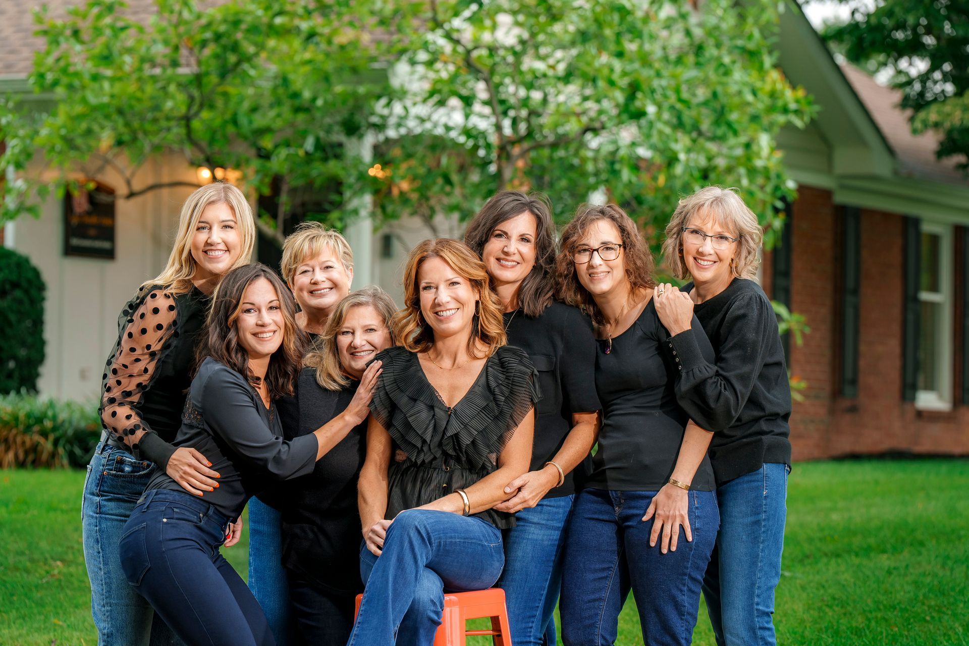 Seven women in teal and white tops posing together outdoors in front of a building and greenery