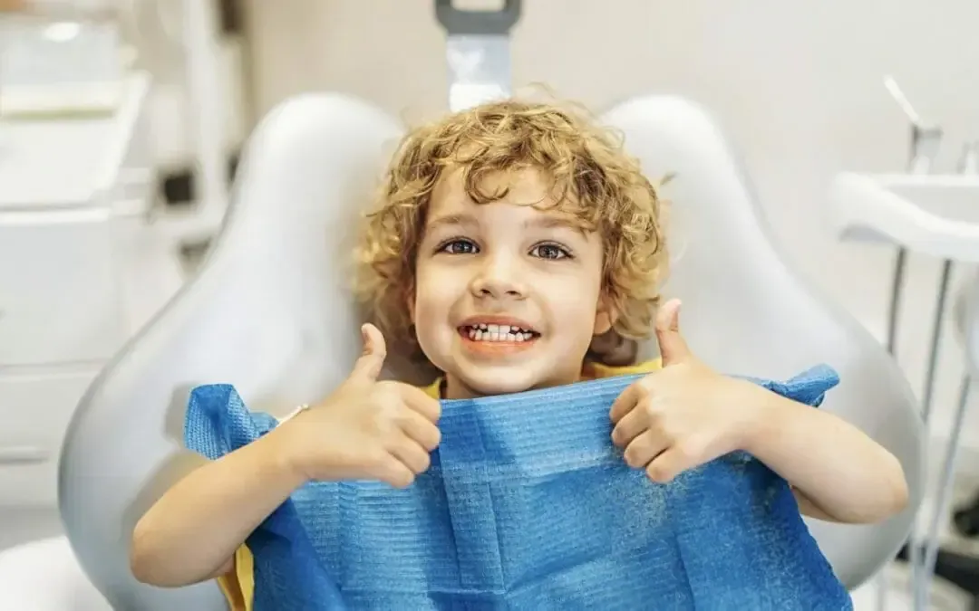 Smiling child in a dental chair giving two thumbs up, holding a blue bib