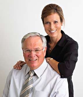 Two smiling coworkers posing in a studio, one seated and one standing behind with a hand on his shoulder.