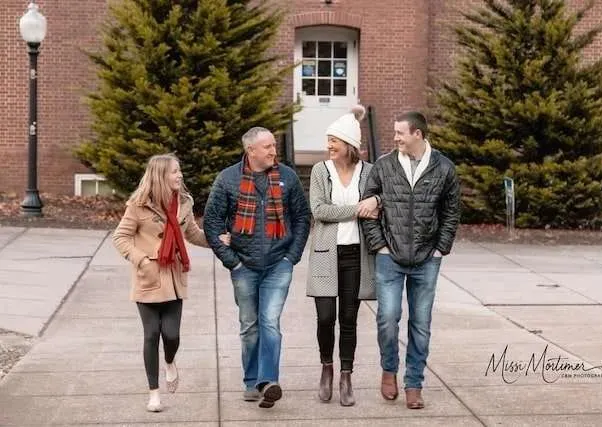 Four people walking together outdoors in front of a brick building and evergreen trees.