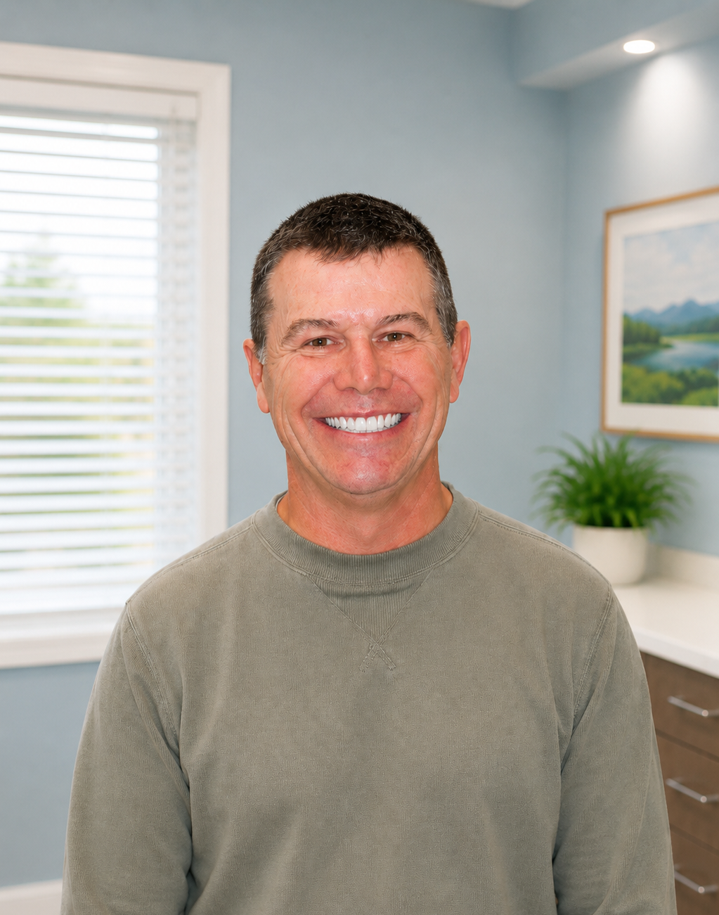 Smiling man in a light green shirt in a bright office, with a window, desk, plant, and framed artwork.