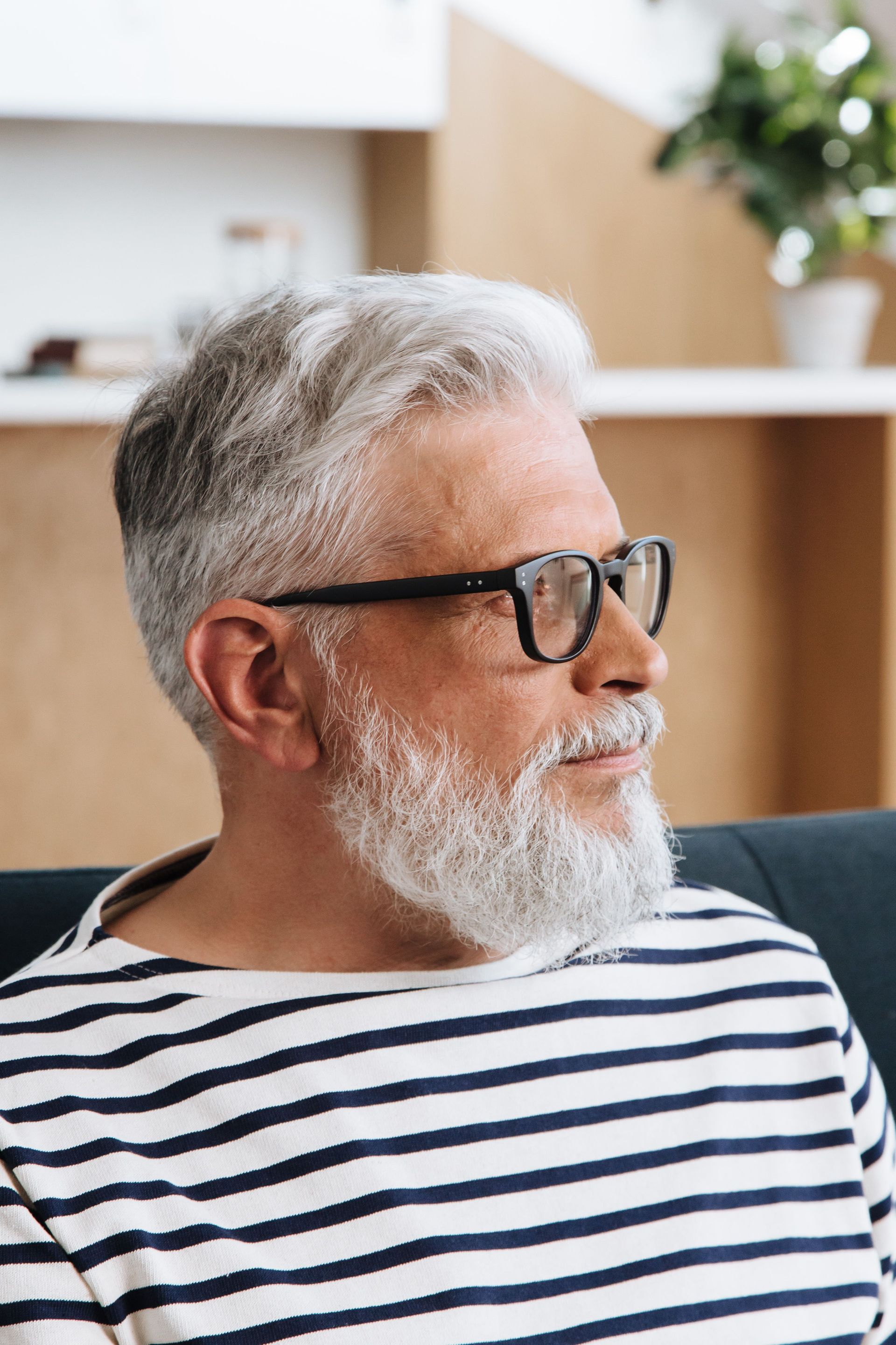 White-bearded man in black glasses and striped shirt, seated indoors on a couch, looking to the side