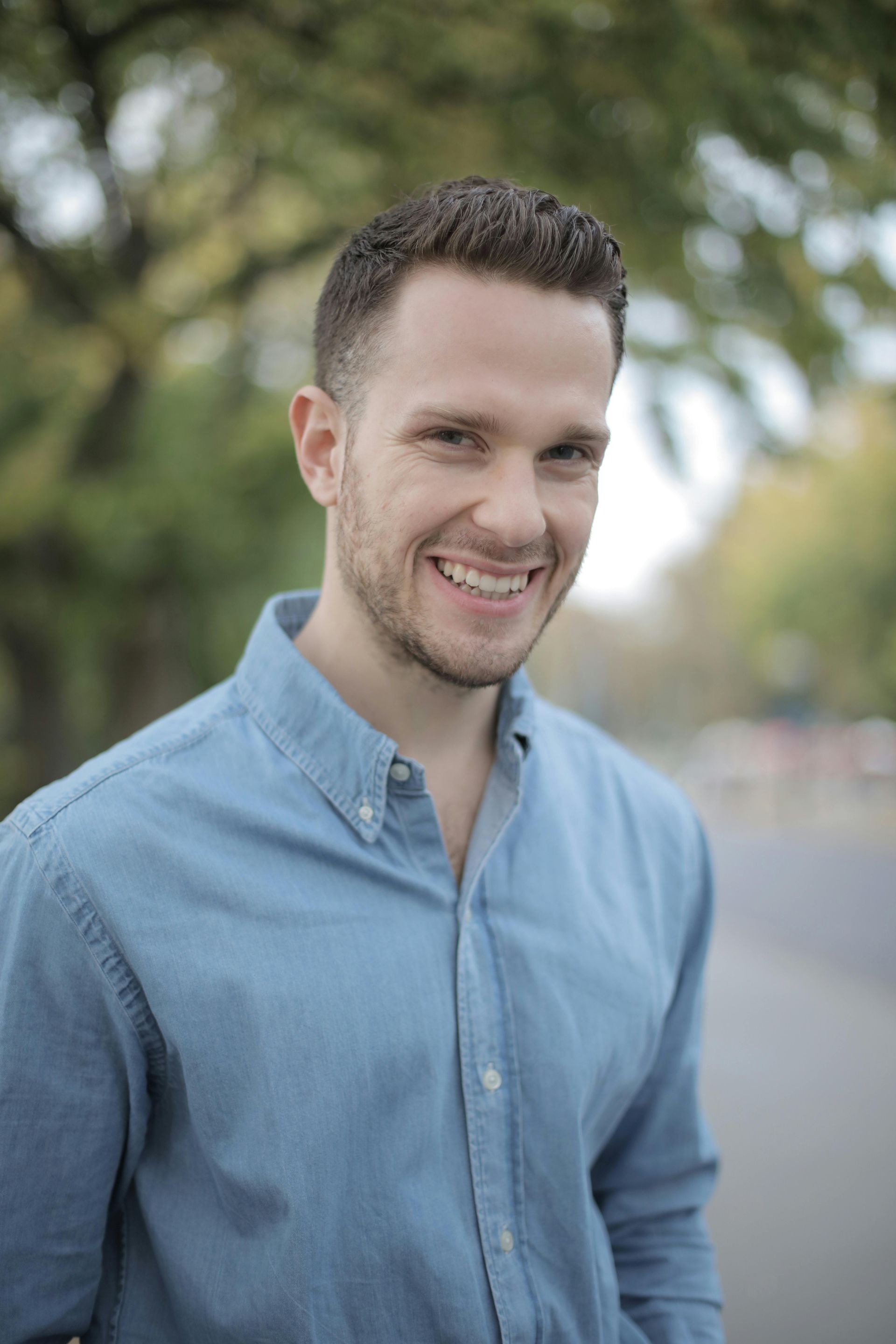 Smiling person in a light blue shirt standing outdoors with blurred trees in the background