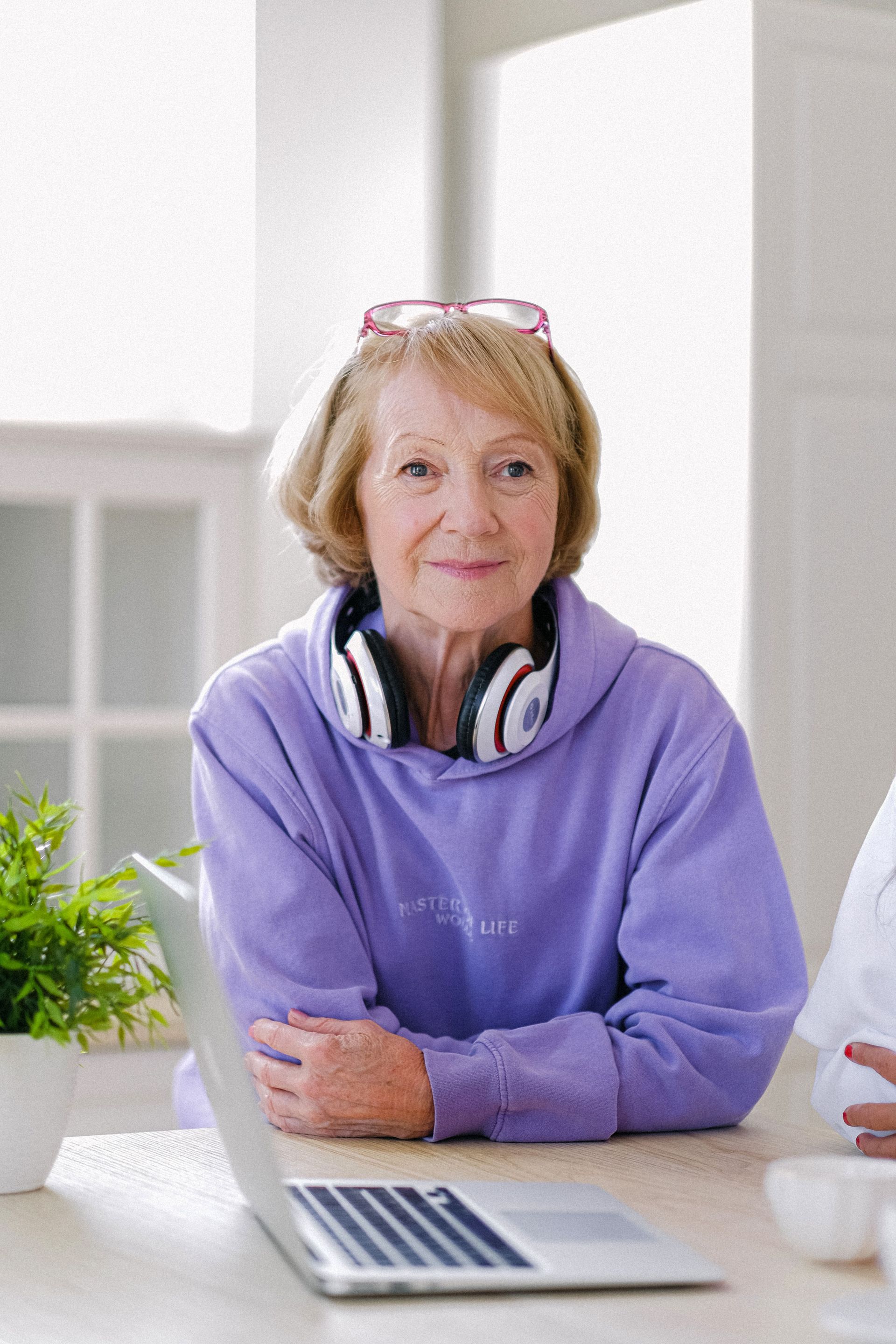 Woman in a purple sweater with headphones around her neck, leaning over a laptop at a bright table.
