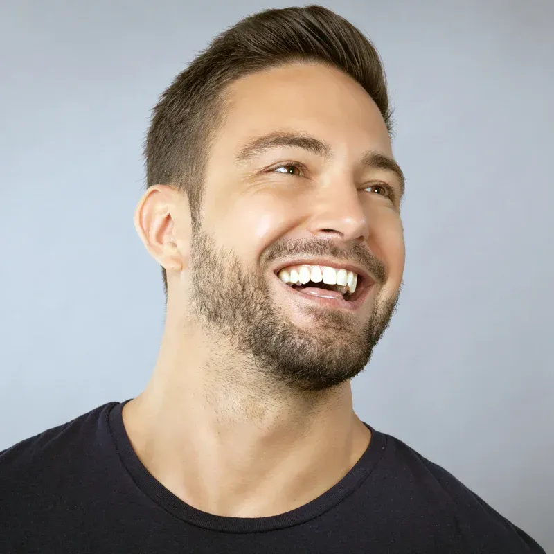 Smiling man with short dark hair and beard, looking to the side against a light blue background