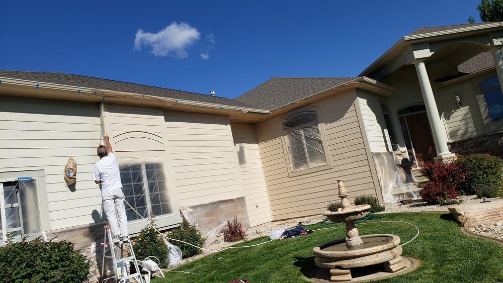 Person painting a beige house exterior beside a green lawn and stone birdbath under a blue sky