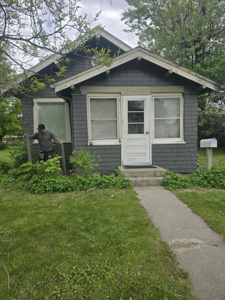 Small gray house with white trim, front porch, and a person standing by the left side yard.