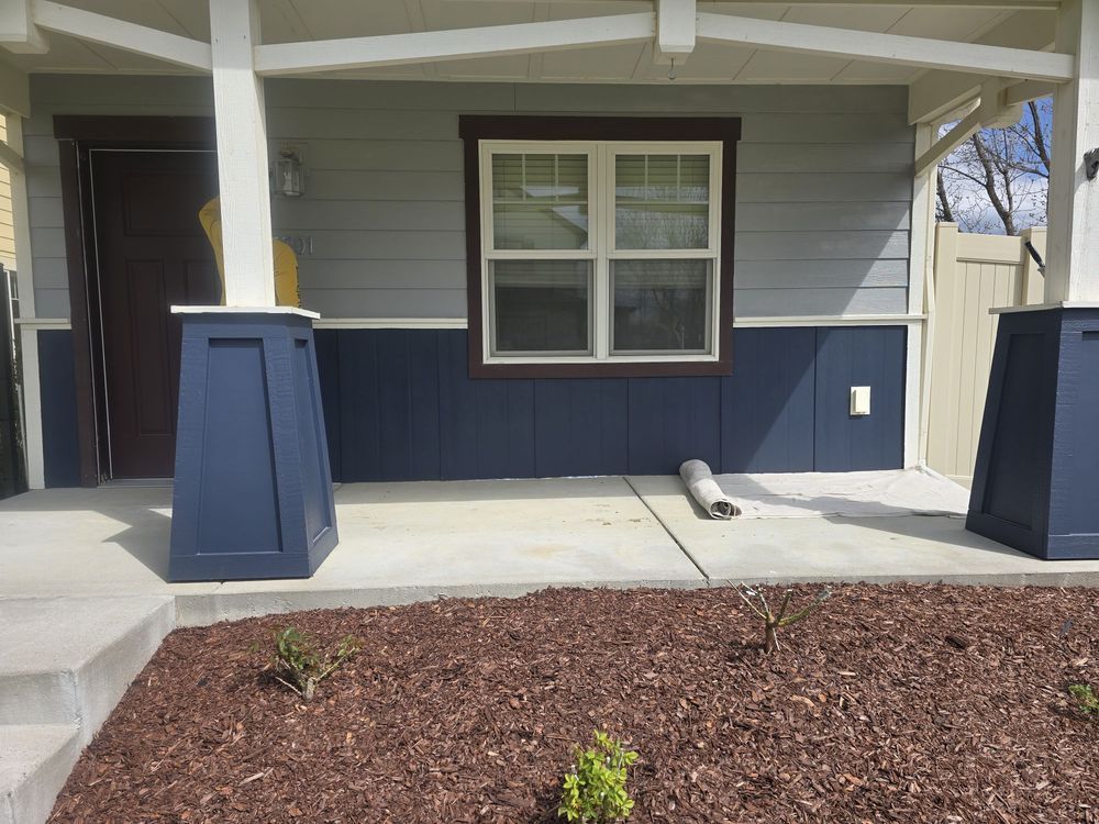 Front porch with blue-gray siding, white columns, dark mulch bed, and a central window above the entry.