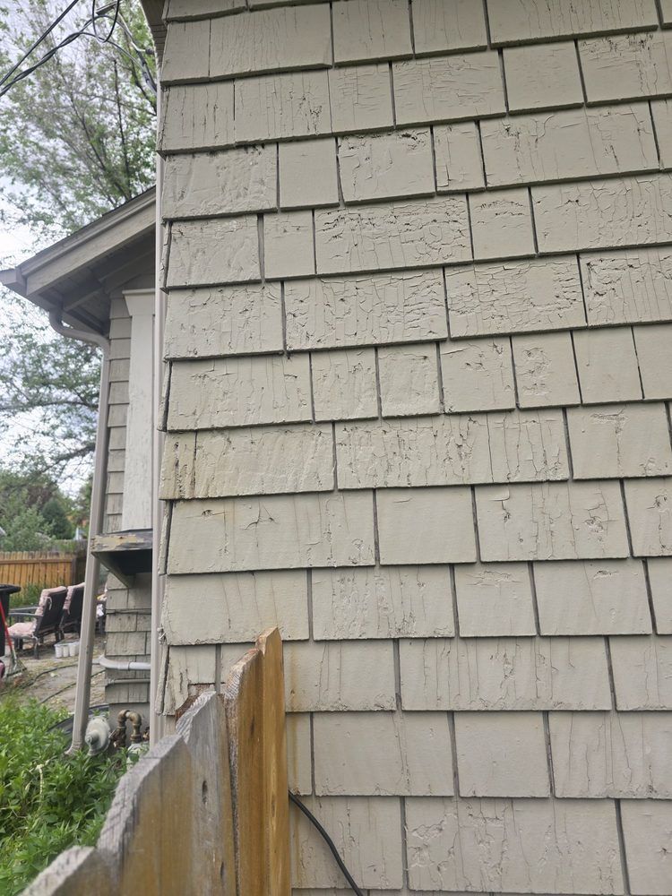 Close-up of gray shingle siding on a house exterior beside a wooden fence and yard.