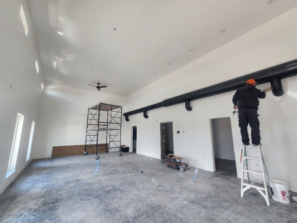 Large white room under renovation with scaffolding, ladders, and a worker installing a black ceiling rail