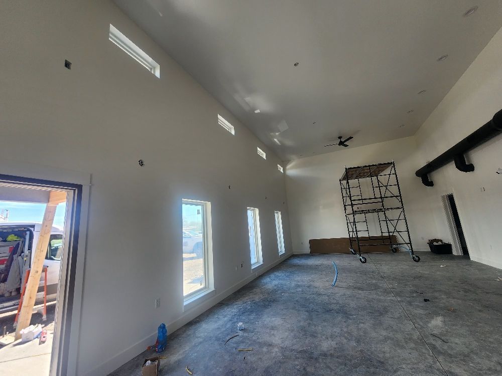 Empty room under renovation with white walls, concrete floor, windows, and scaffolding near the back wall