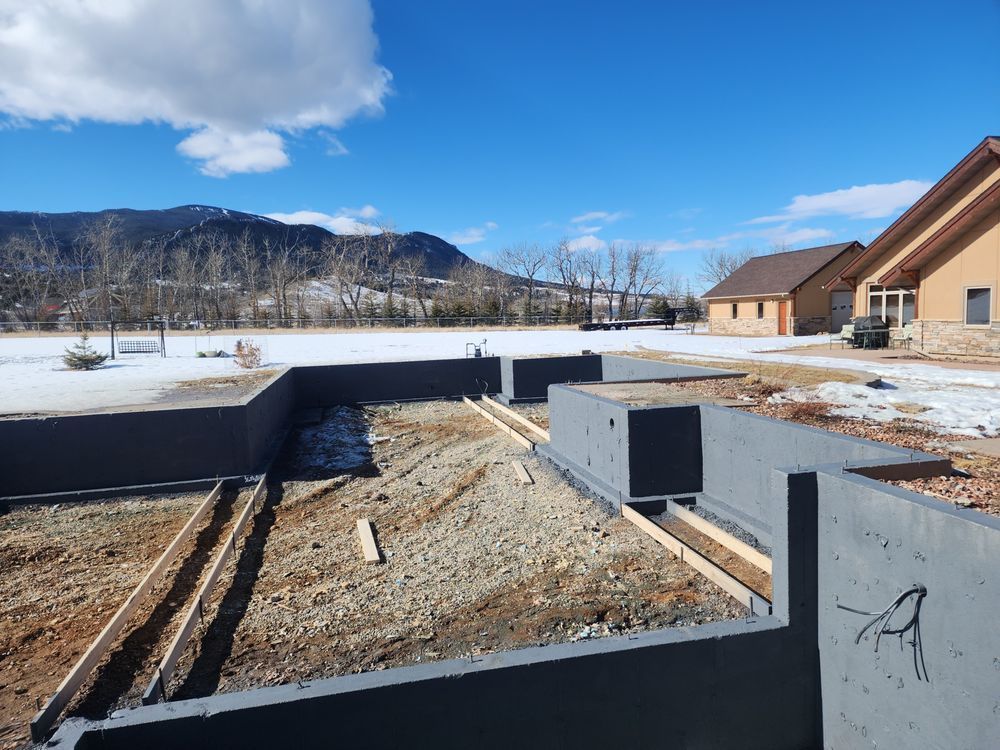Foundation walls under construction beside a snowy rural neighborhood with mountains in the distance