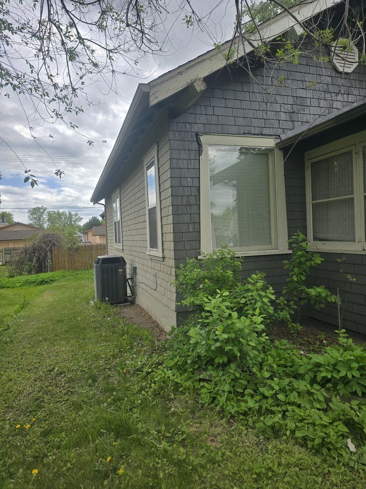 Side of a gray house with a bay window, overgrown grass, and cloudy sky.