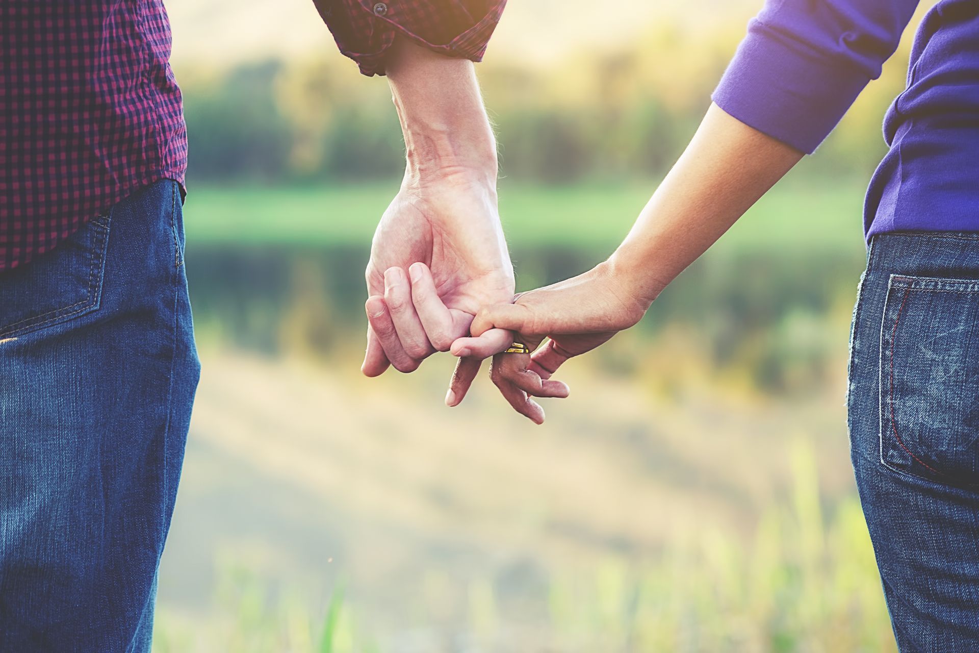 Two people holding hands outdoors in warm sunlight near a blurred path and greenery