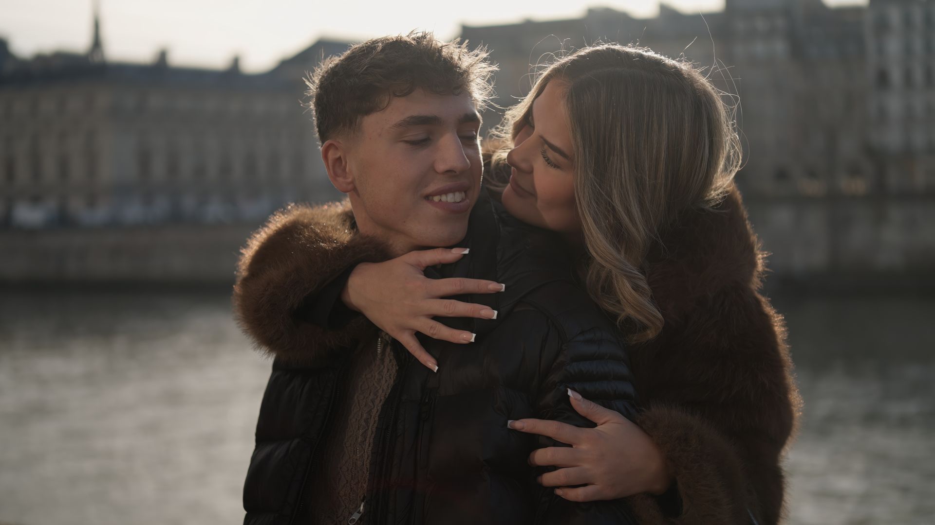 Two people embracing by a waterfront at sunset, with a city building in the background.
