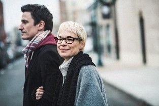 Two people standing arm in arm on a city street, one wearing glasses and smiling at the camera.