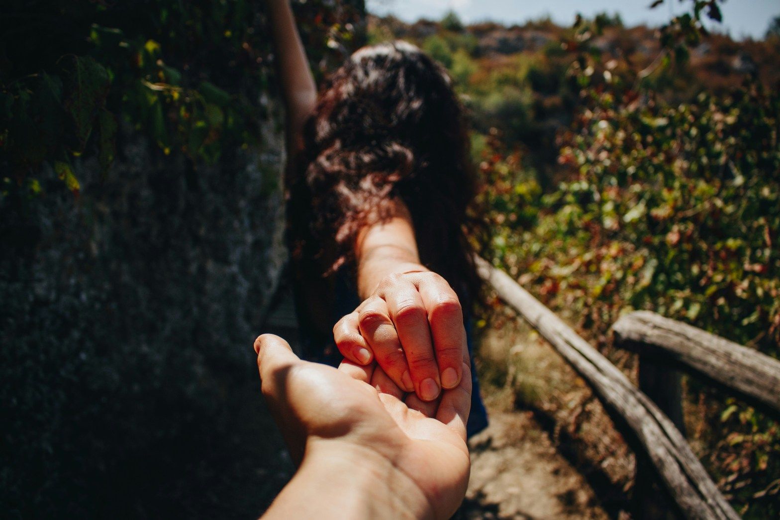 Hand reaching for another hand on a sunlit outdoor trail near greenery and a wooden rail