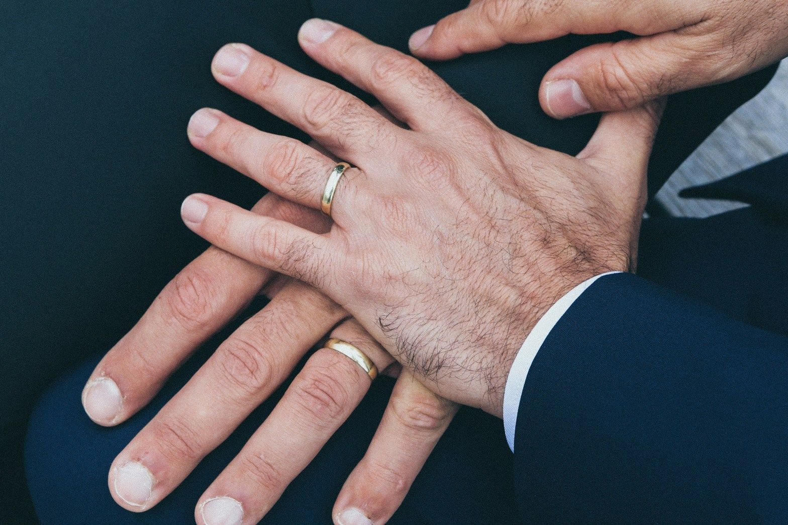 Hands stacked together in a team gesture, showing wedding rings and dark suit sleeves.