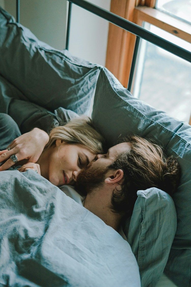 Couple sleeping together in bed under a teal blanket by a window