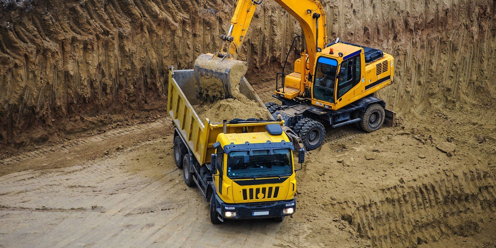 Un escavatore giallo carica un camion con cassone ribaltabile giallo con del terreno in un cantiere edile.