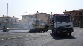 Attrezzatura per la pavimentazione stradale e camion su una strada, con edifici sullo sfondo sotto un cielo luminoso.