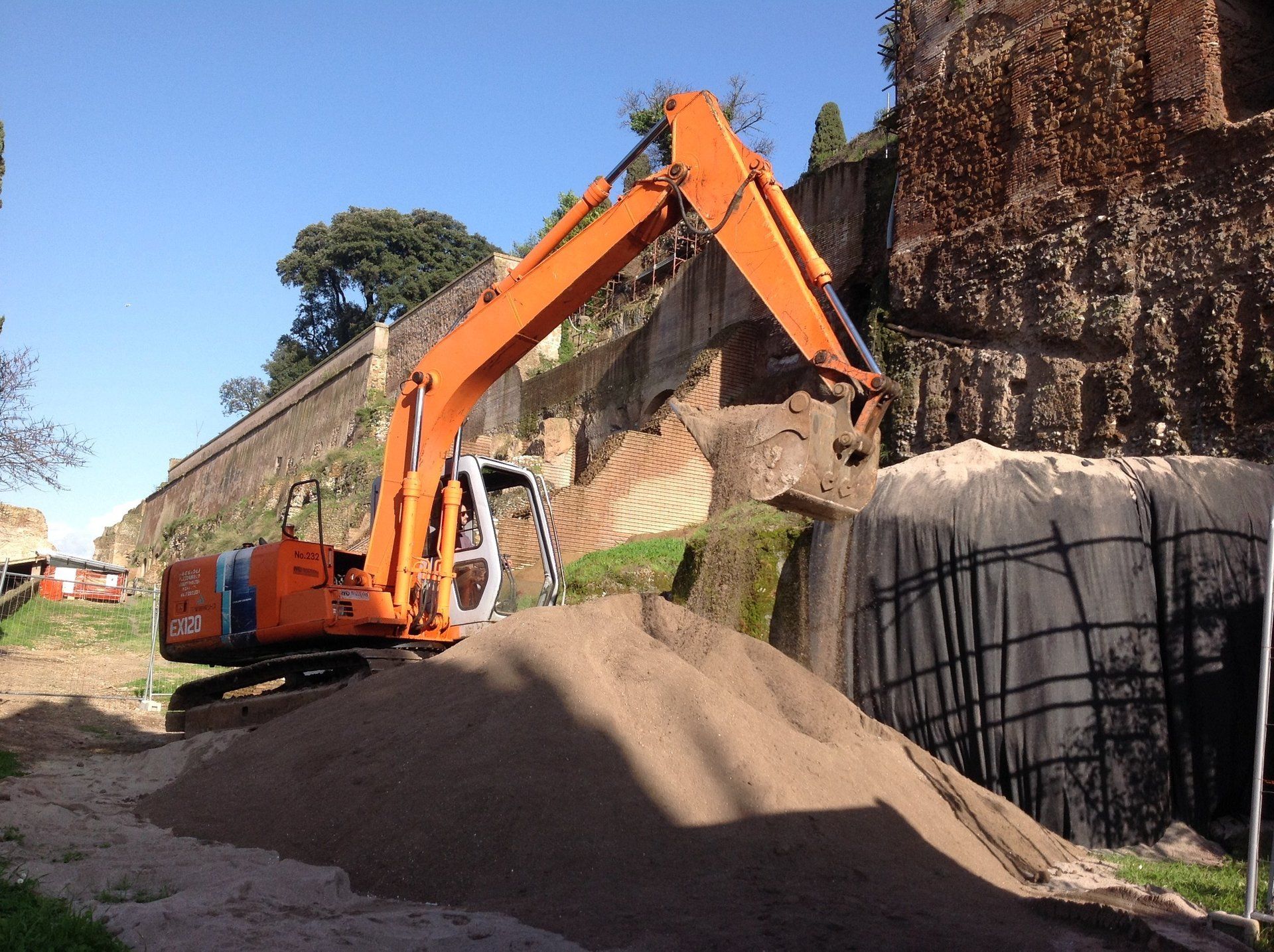 Escavatore arancione che sposta sabbia vicino a un muro di pietra, probabilmente per lavori di costruzione o scavo.
