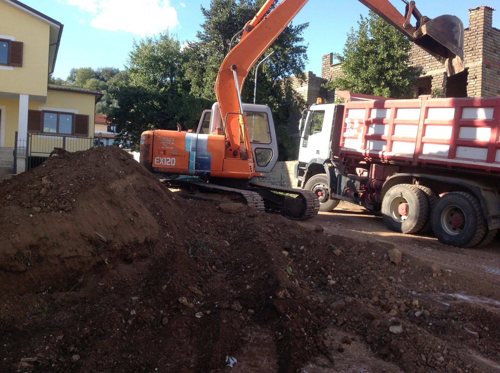 Un escavatore arancione carica un camion con cassone ribaltabile pieno di terra in un cantiere edile, vicino a edifici e alberi.