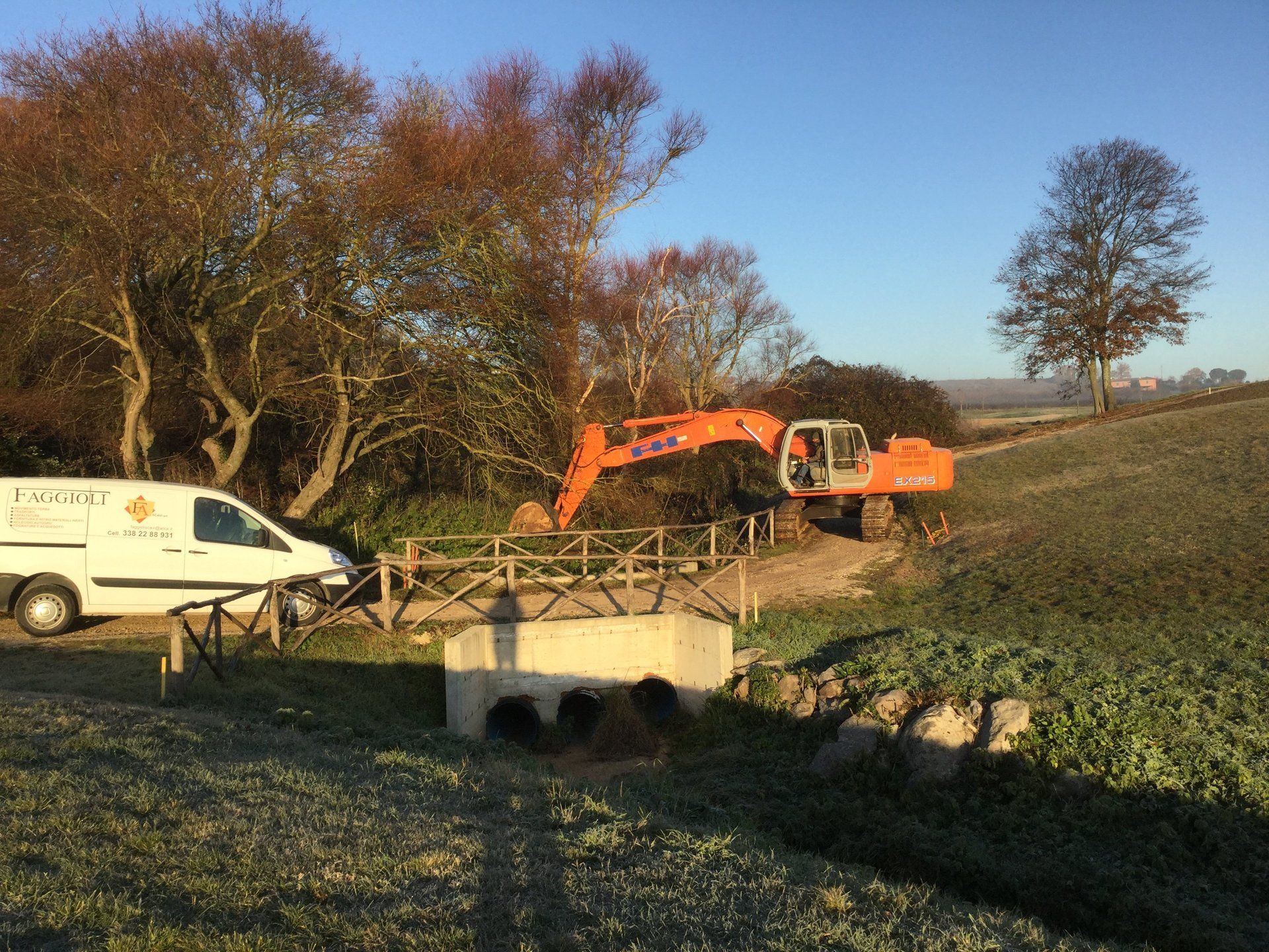 Escavatore al lavoro vicino a un piccolo ponte in un campo. Furgone bianco parcheggiato lì vicino. Giornata di sole.