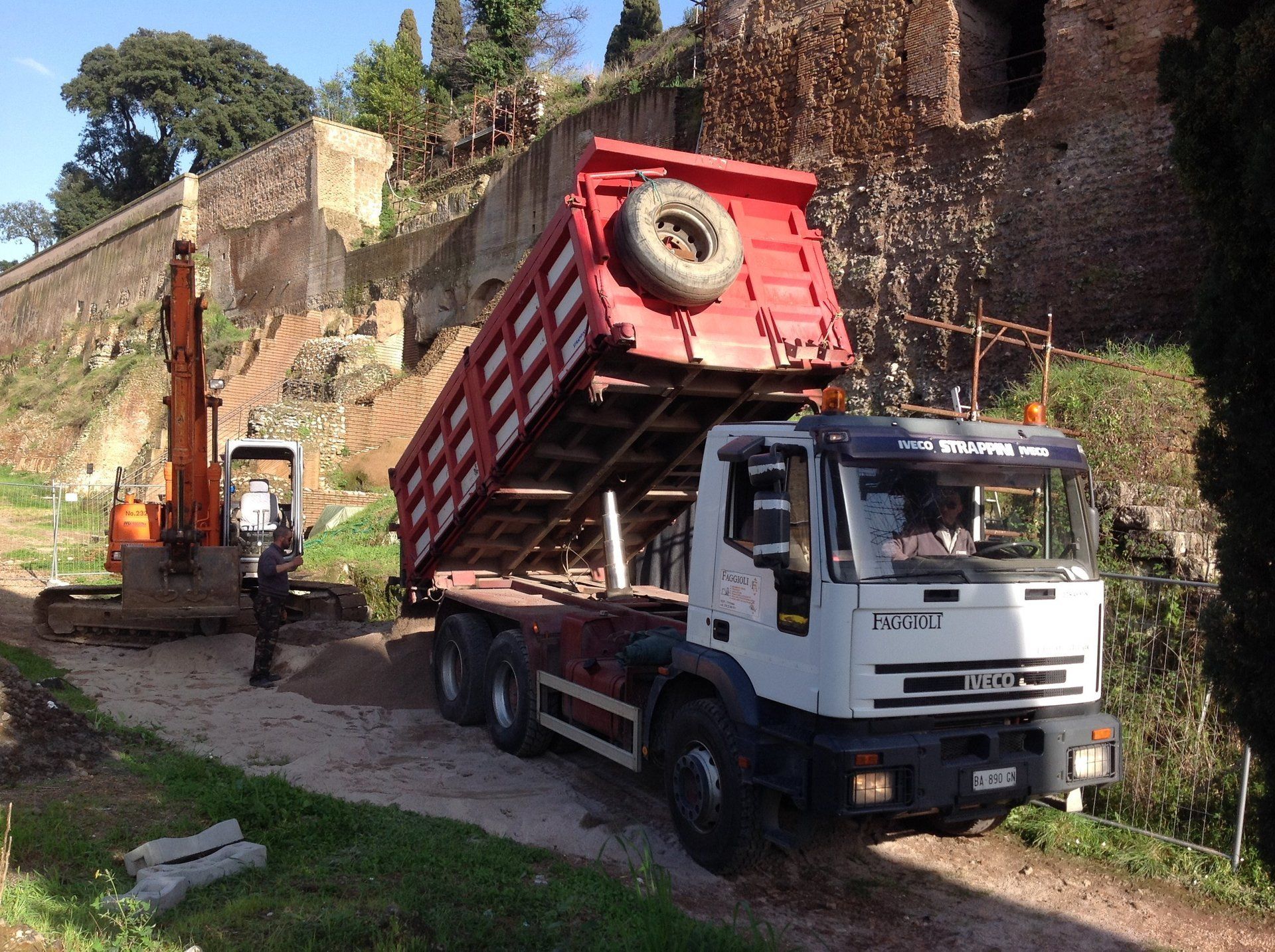 Autocarro con cassone ribaltabile che scarica materiale accanto a un escavatore e a un antico muro di pietra.