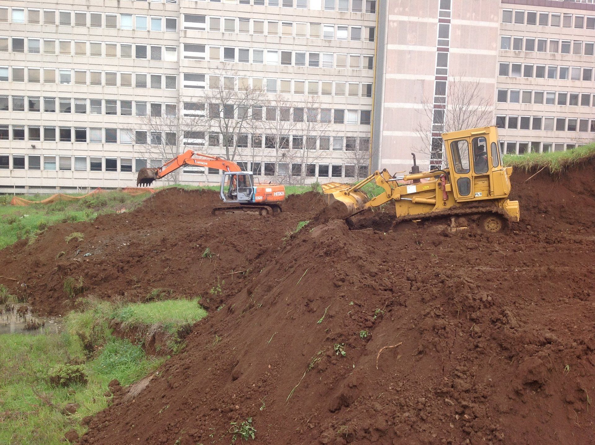 Cantiere con una ruspa e un escavatore che spostano terra vicino a un edificio alto.