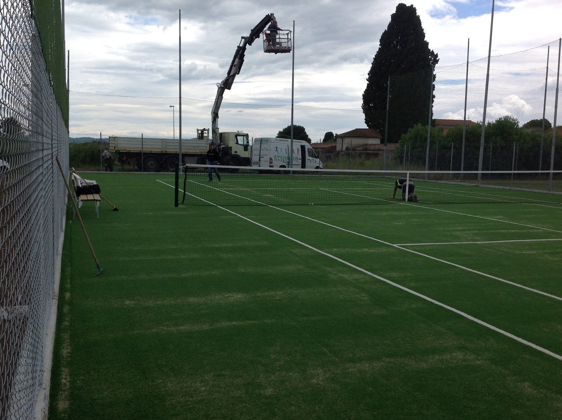 Campo da tennis con piattaforma di lavoro elevata che installa i pali della rete sotto un cielo coperto.