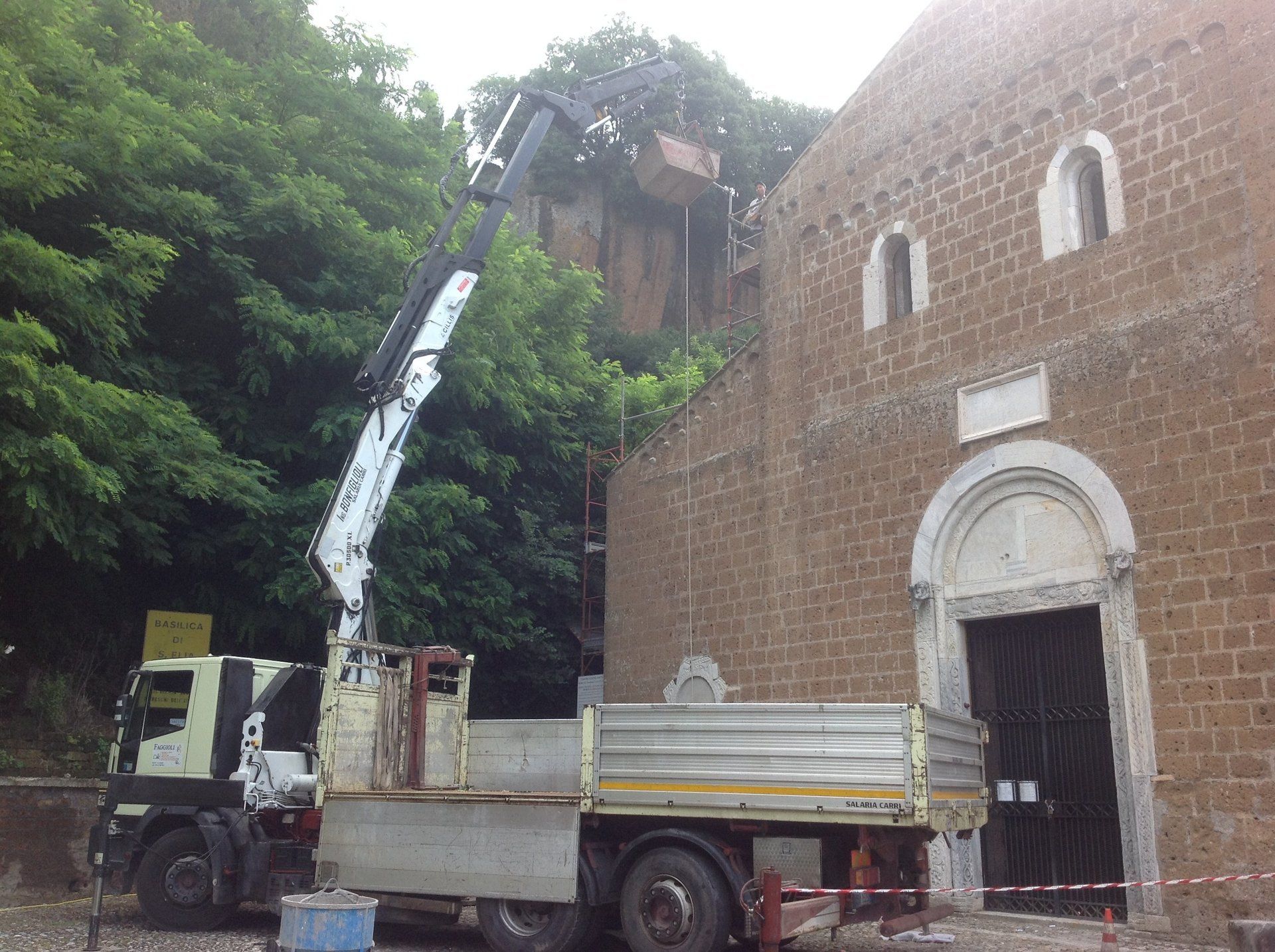 Camion con gru accanto a una chiesa in pietra, probabilmente per lavori di costruzione o manutenzione. Alberi sullo sfondo.