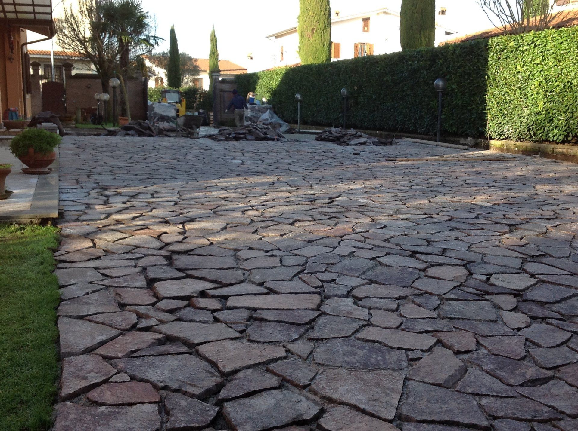 Area esterna pavimentata in pietra con siepe verde e alberi.