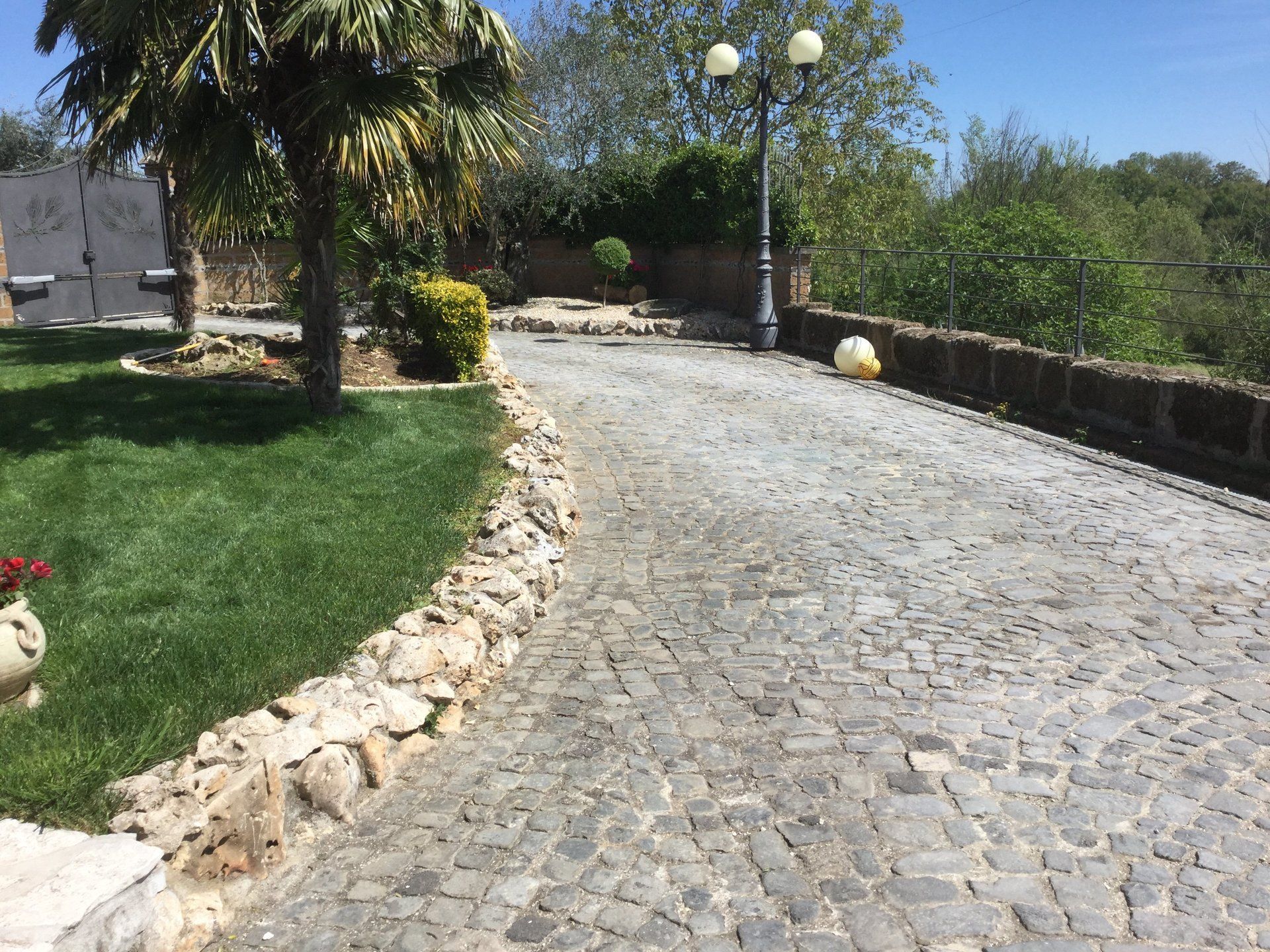 Vialetto di ciottoli che costeggia un prato verde, bordato da pietre, e che conduce a un giardino con un lampione.