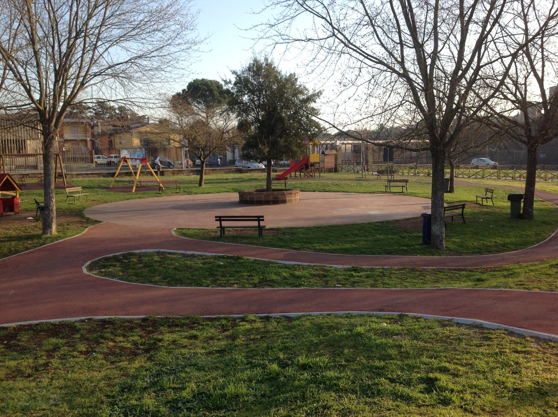 Parco con sentiero pedonale rosso, parco giochi, alberi, panchine ed erba.