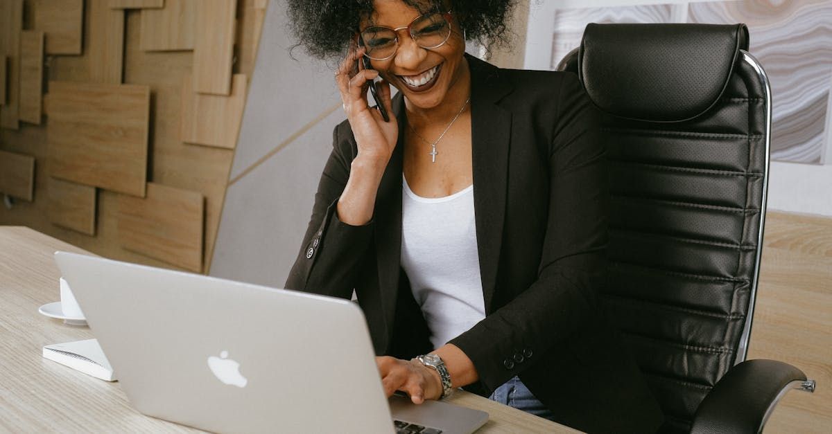 A woman is sitting at a desk using a laptop and talking on a cell phone.