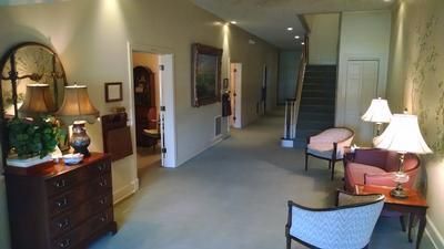 Long hallway in a home or lobby with chairs, lamps, stairs, and framed wall art.